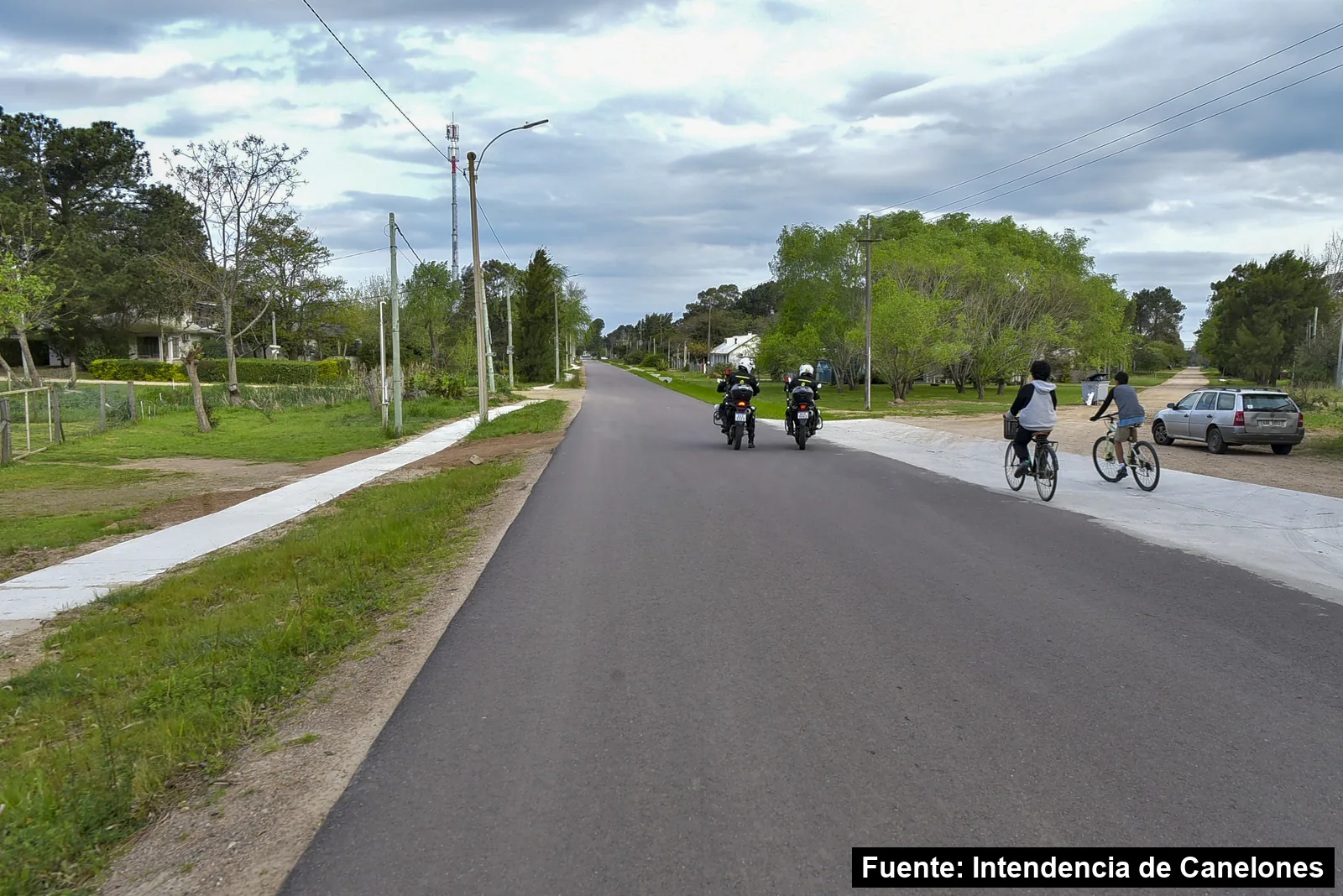 Calle recién pavimentada con senda peatonal de hormigón, dos policías en moto y dos jóvenes en bicicleta en el Municipio de La Floresta