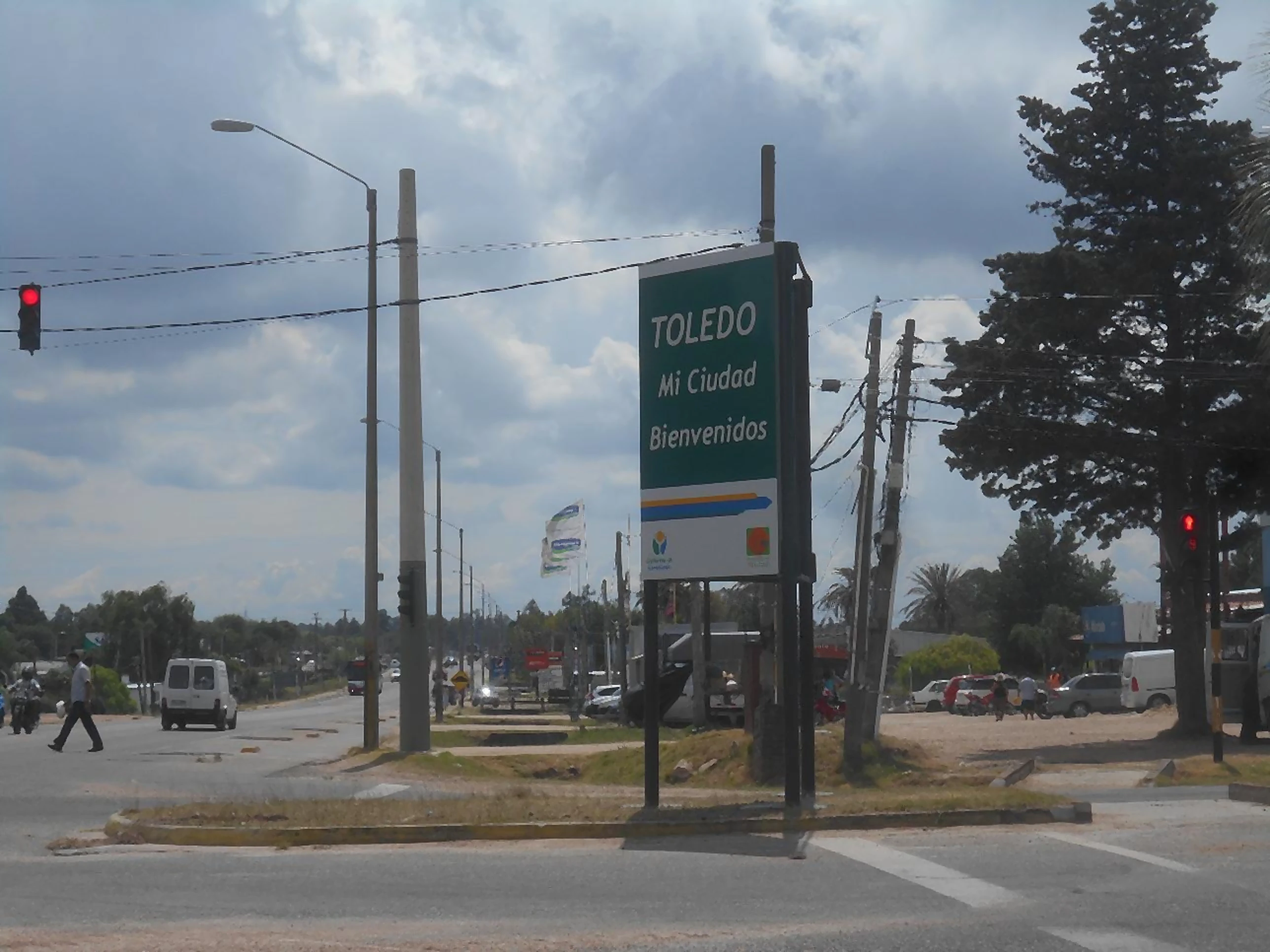 Mayo Amarillo en Toledo 1 Cartel verde de bienvenida a Toledo junto al cruce de dos rutas, con semáforos y tránsito, Canelones