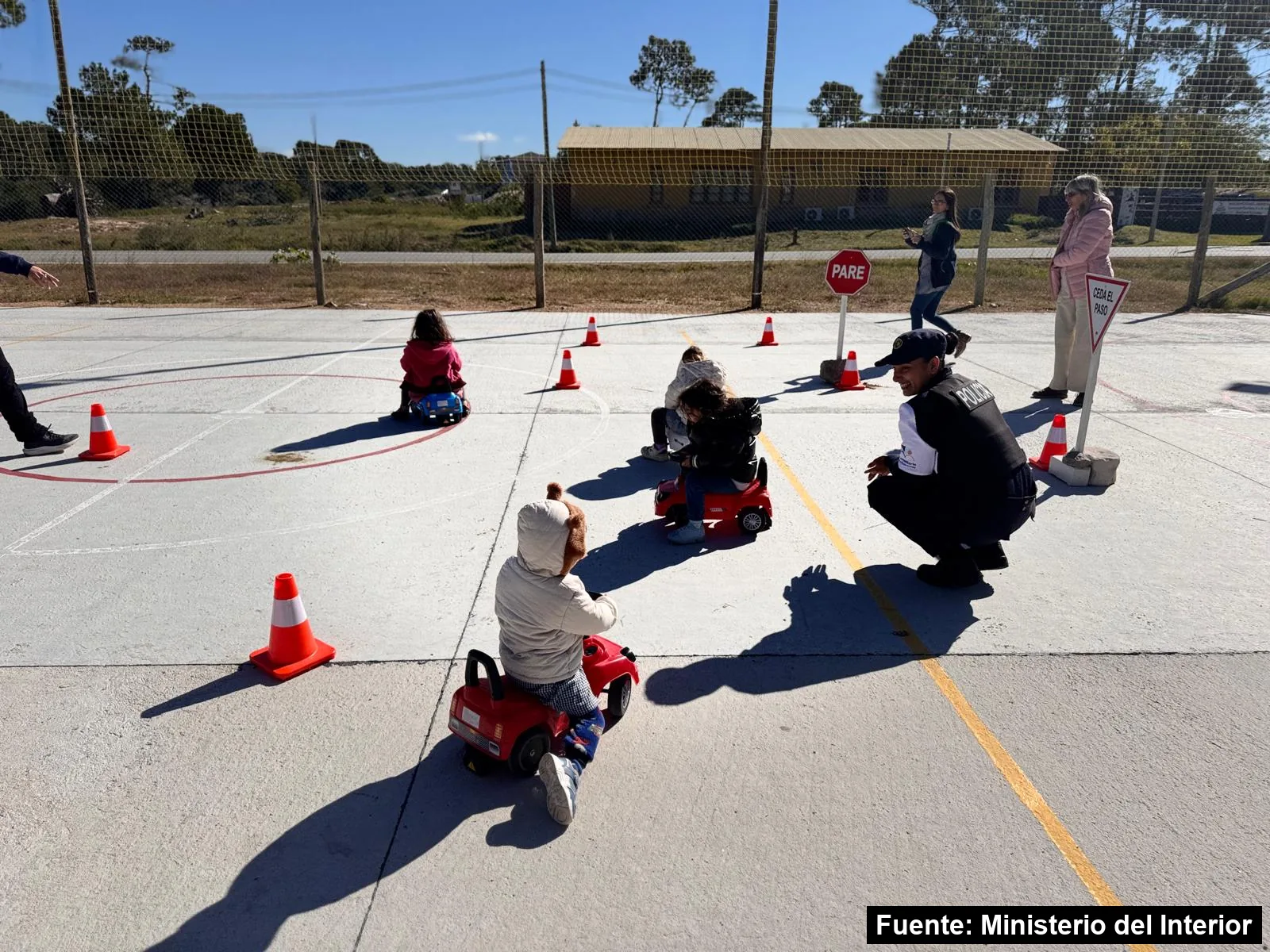 Mayo Amarillo en La Paloma 4 Niños pequeños de espaldas en autitos de juguete recorren un circuito con conos y señales de Pare y Ceda el paso en una cancha, junto a un policía agachado