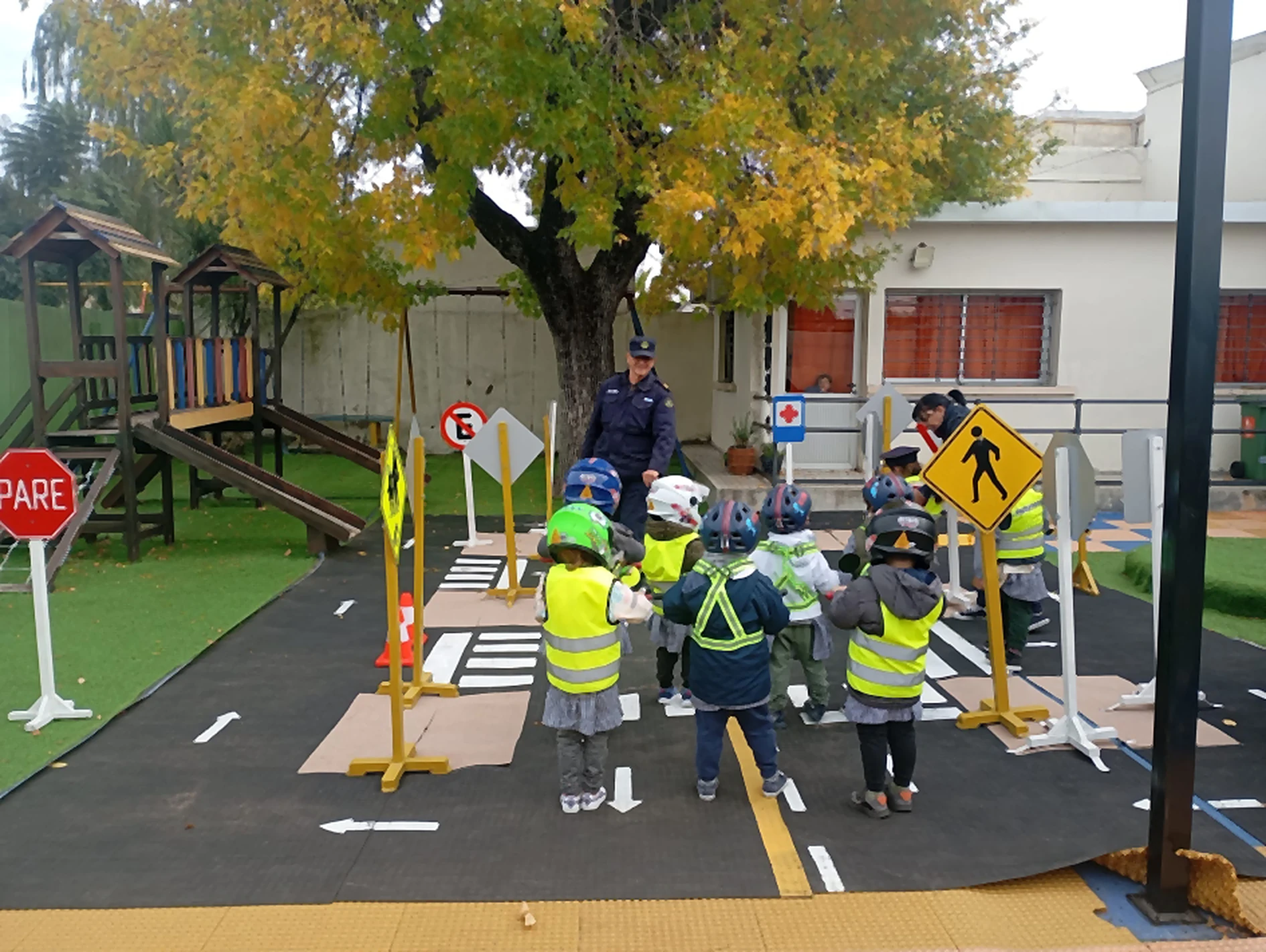 Niños de espaldas, con casco y chaleco reflectivo, recorren una calle armada en el patio de un jardín, con señales de tránsito y un policía al fondo