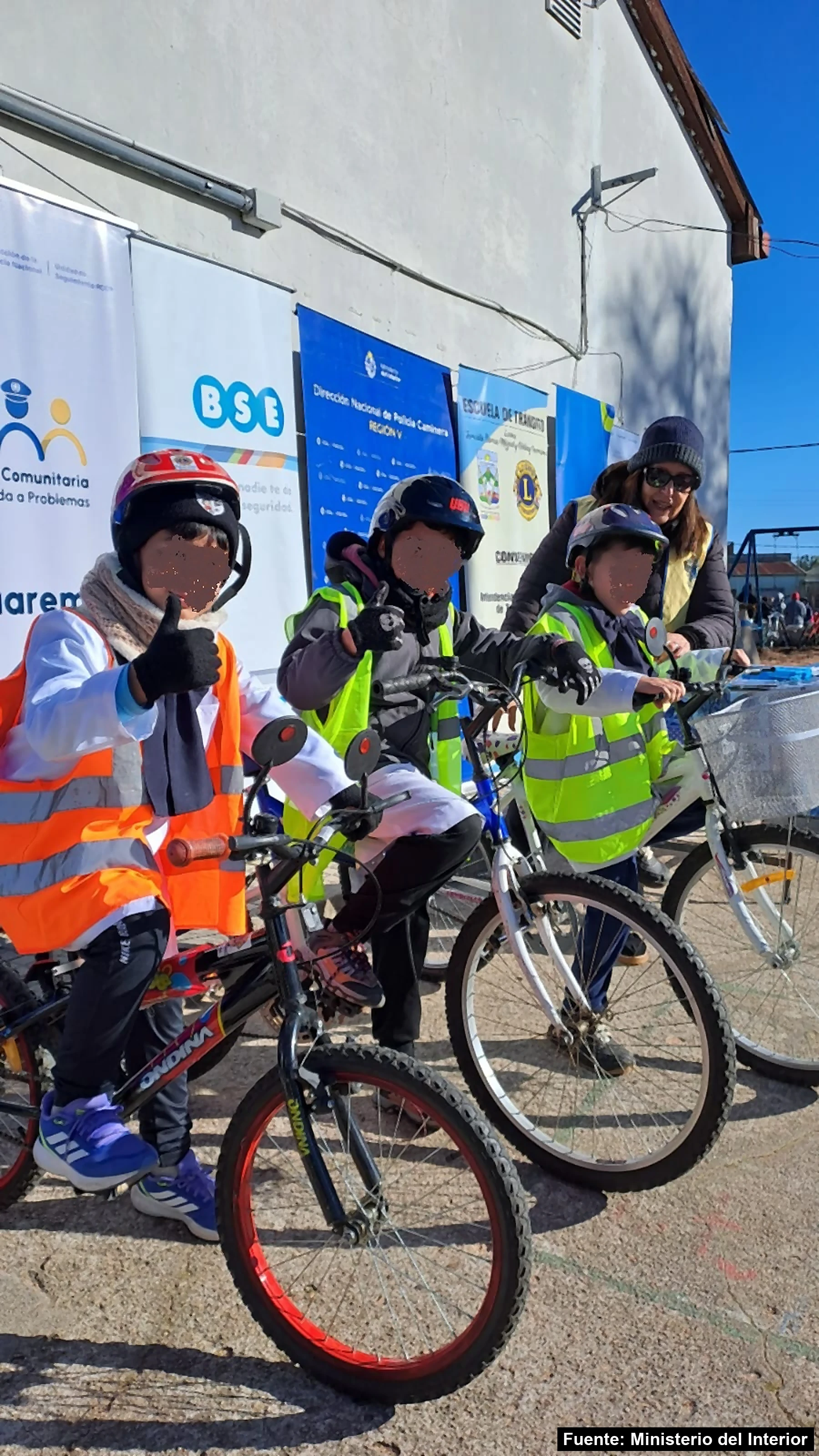 Escolares con casco y chaleco reflectivo en bicicleta frente a los carteles de la Policía Comunitaria, el BSE, la Policía Caminera y la Escuela de Tránsito, en Ansina