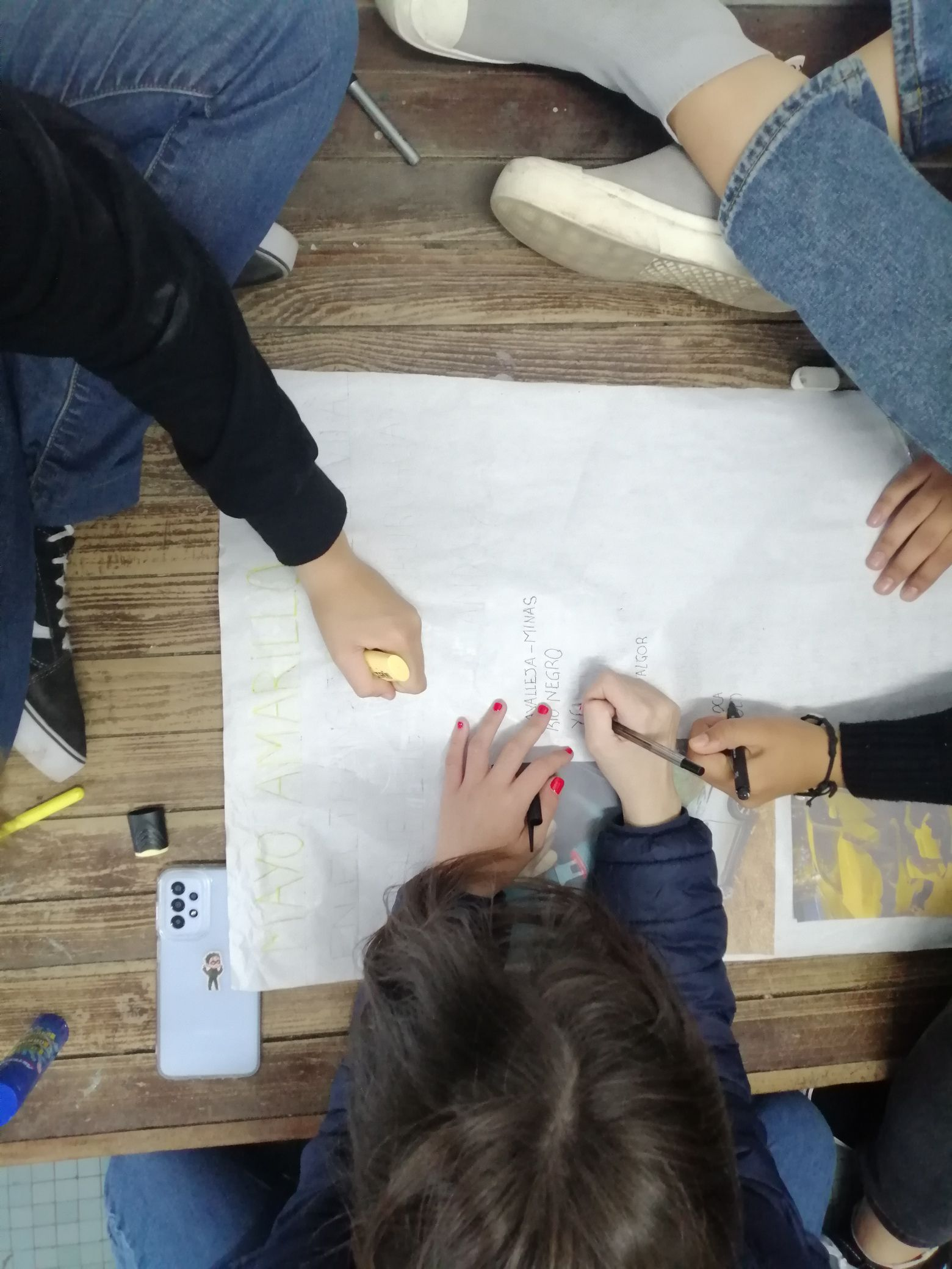 Manos de estudiantes escribiendo y pintando una cartelera de Mayo Amarillo sobre una mesa de madera, vista desde arriba
