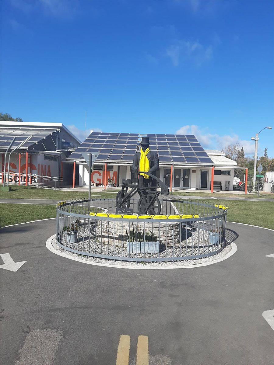 Monumento a Karl Drais con una bufanda amarilla en la rotonda del circuito vial de la Plaza de Deportes de Canelones
