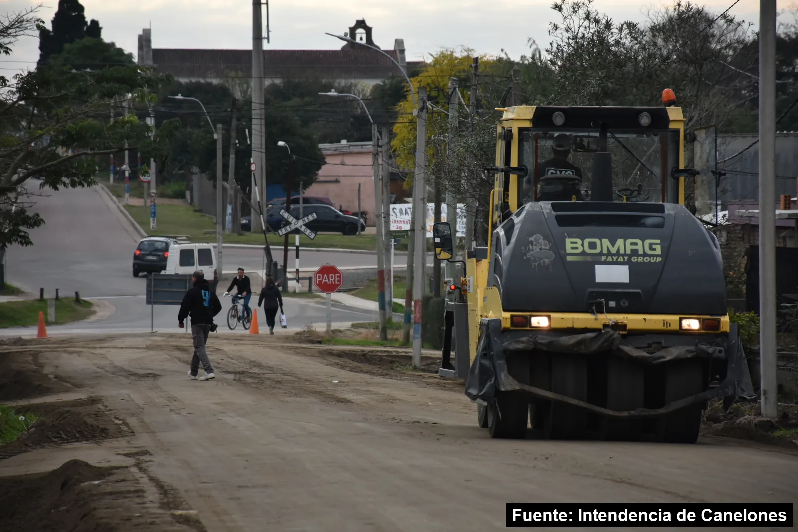 Compactadora trabajando en una calle de tierra de Joaquín Suárez mientras pasan peatones y un ciclista