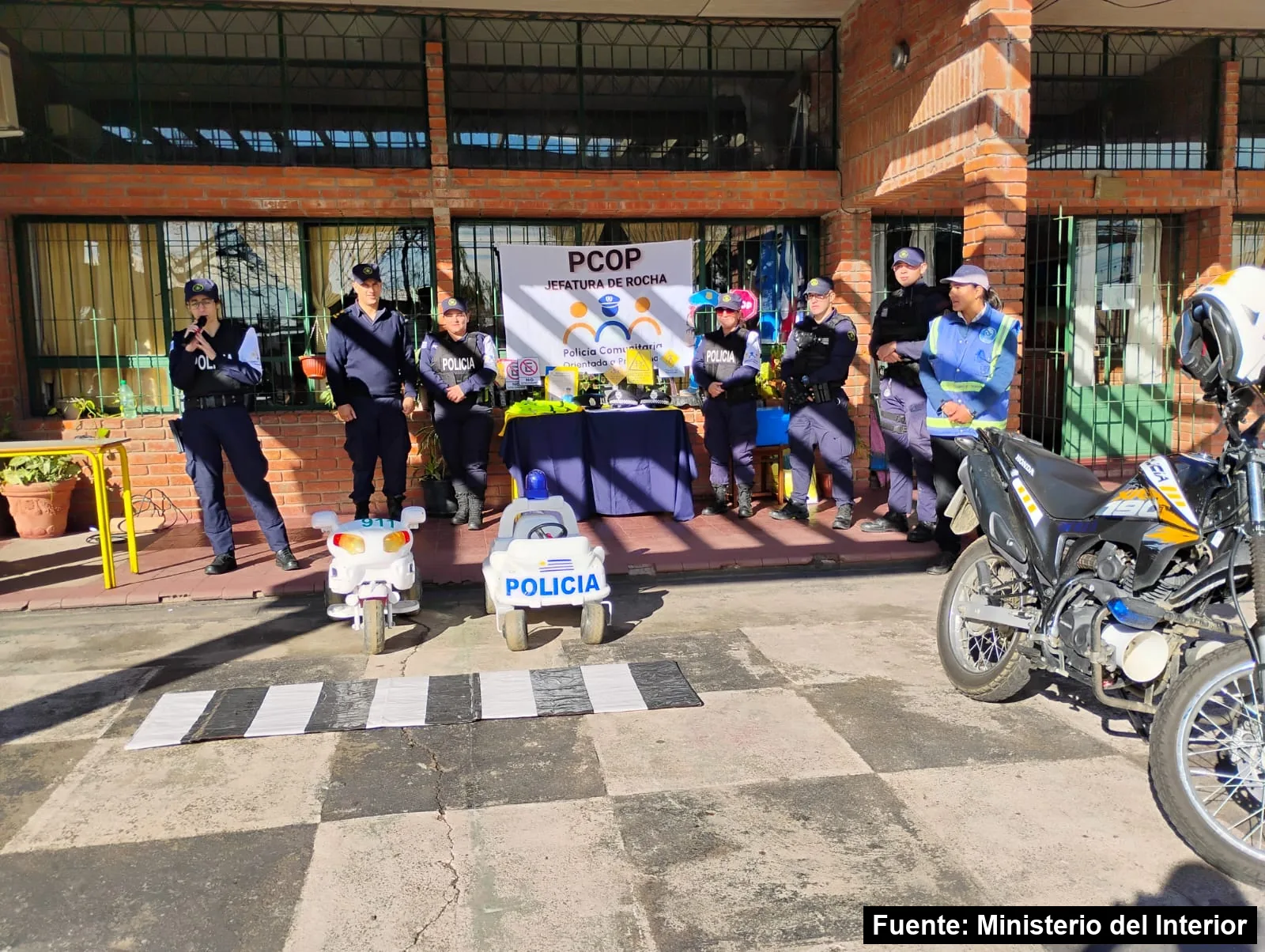 Policías de la Policía Comunitaria y un inspector de tránsito frente a una escuela de ladrillo, con una senda peatonal, un auto y una moto de juguete y una moto policial