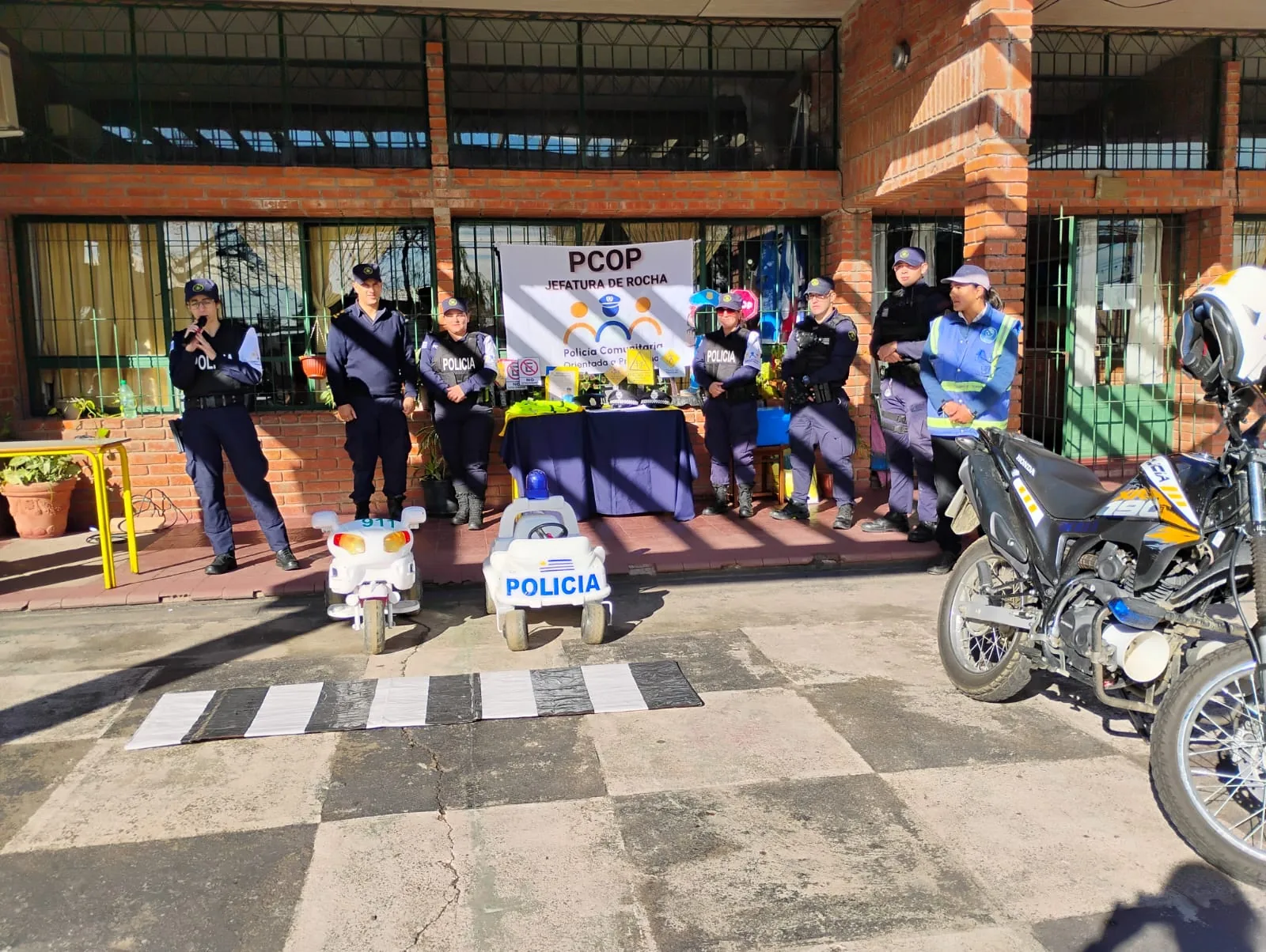 Mayo Amarillo en Lascano 1 Policías de la Policía Comunitaria y un inspector de tránsito frente a una escuela de ladrillo, con una senda peatonal, un auto y una moto de juguete y una moto policial
