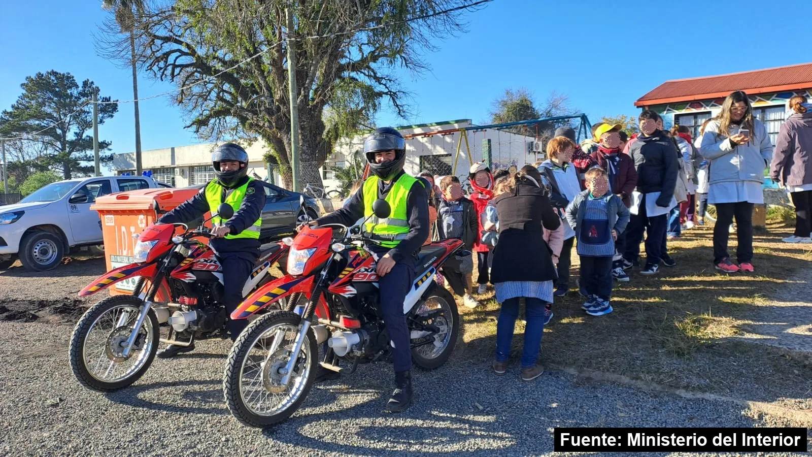 Dos policías con casco y chaleco reflectivo sobre motos rojas, junto a una fila de escolares en el patio de una escuela de Ansina