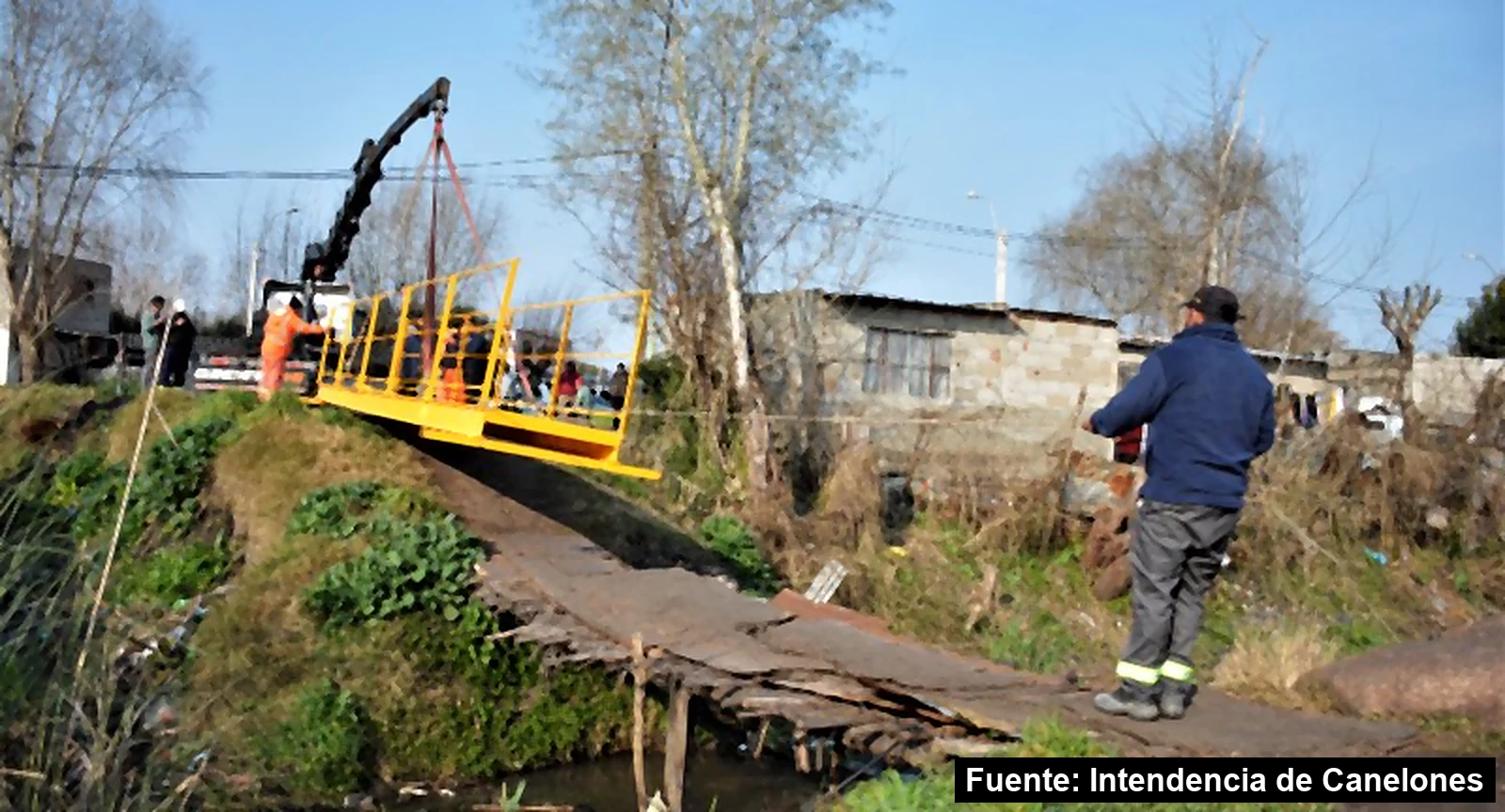 Grúa colocando un puente peatonal de acero con barandas amarillas sobre una cañada en 18 de Mayo
